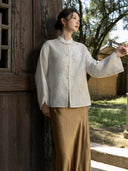 Model raising one arm to show the wide sleeve and split cuff of an ivory jacket with floral chest embellishment.