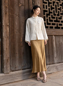 Model standing against a wooden wall in an ivory floral jacquard jacket paired with a long bronze skirt.