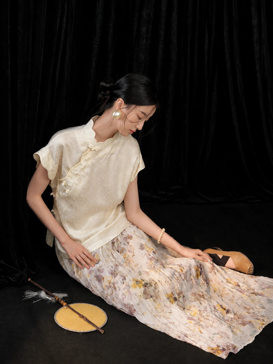 Seated model wearing an ivory mulberry silk short sleeve qipao collar top with a soft floral skirt