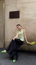 Model sitting on a bench in a sage green sleeveless Qipao top, holding a textured lime green handbag, showcasing the relaxed A-line fit and Lyocell blend fabric.