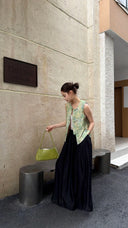 Side view of a woman in a jade floral brocade Cheongsam vest, looking down while holding a bright green bag, highlighting the sleeveless design and silky drape.