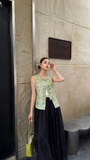 Woman standing by stone wall wearing a jade green Cheongsam vest with watercolor floral brocade, paired with black wide-leg pants and a green bag.