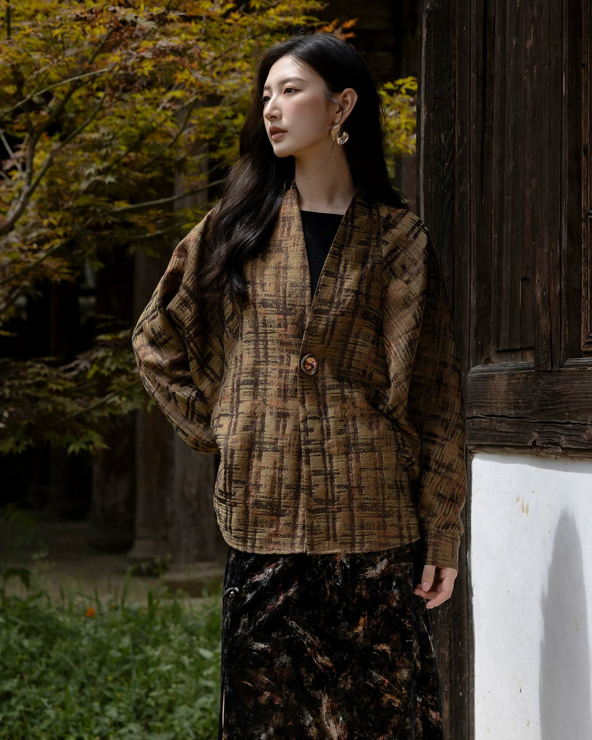 Model wearing the buttoned Mingcha Brown plaid jacket beside a dark wooden wall.