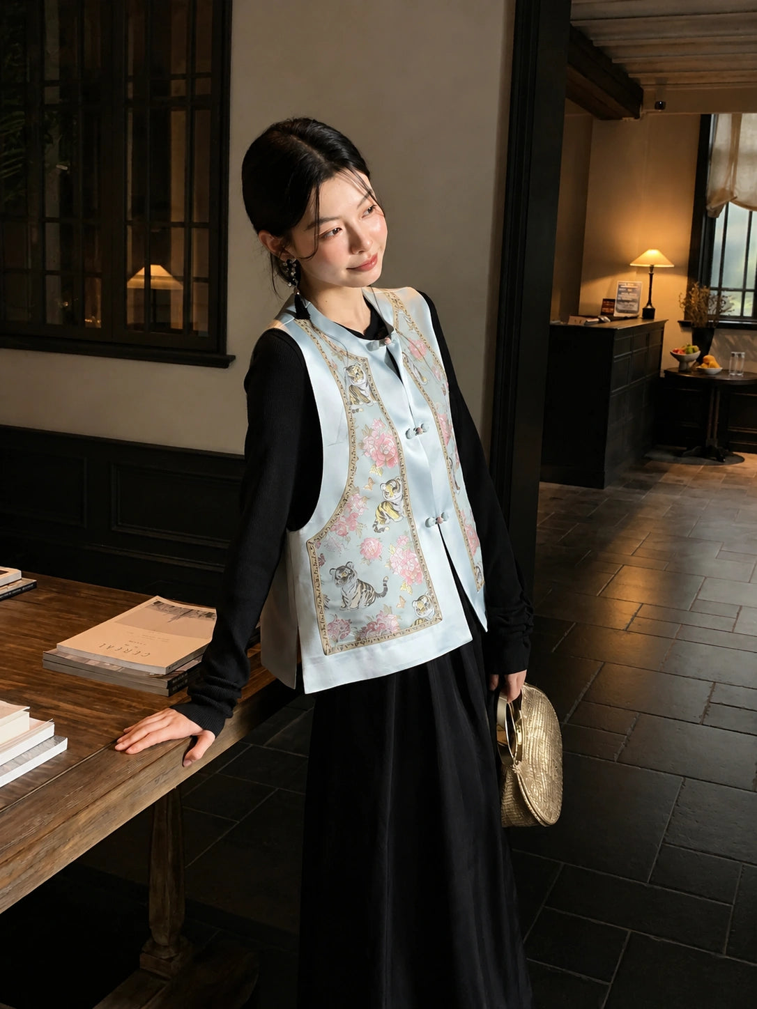 Styling image of a light blue Chinese silk vest with round neckline, floral and tiger pattern, worn over a black dress.