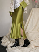 Detail view of a glossy matcha green acetate skirt with a side slit, styled with a white green jacket cuff and black ankle boots.