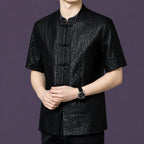 Men's relaxed-fit black silk top with a mandarin collar and frog button closure, styled with black trousers and a watch.