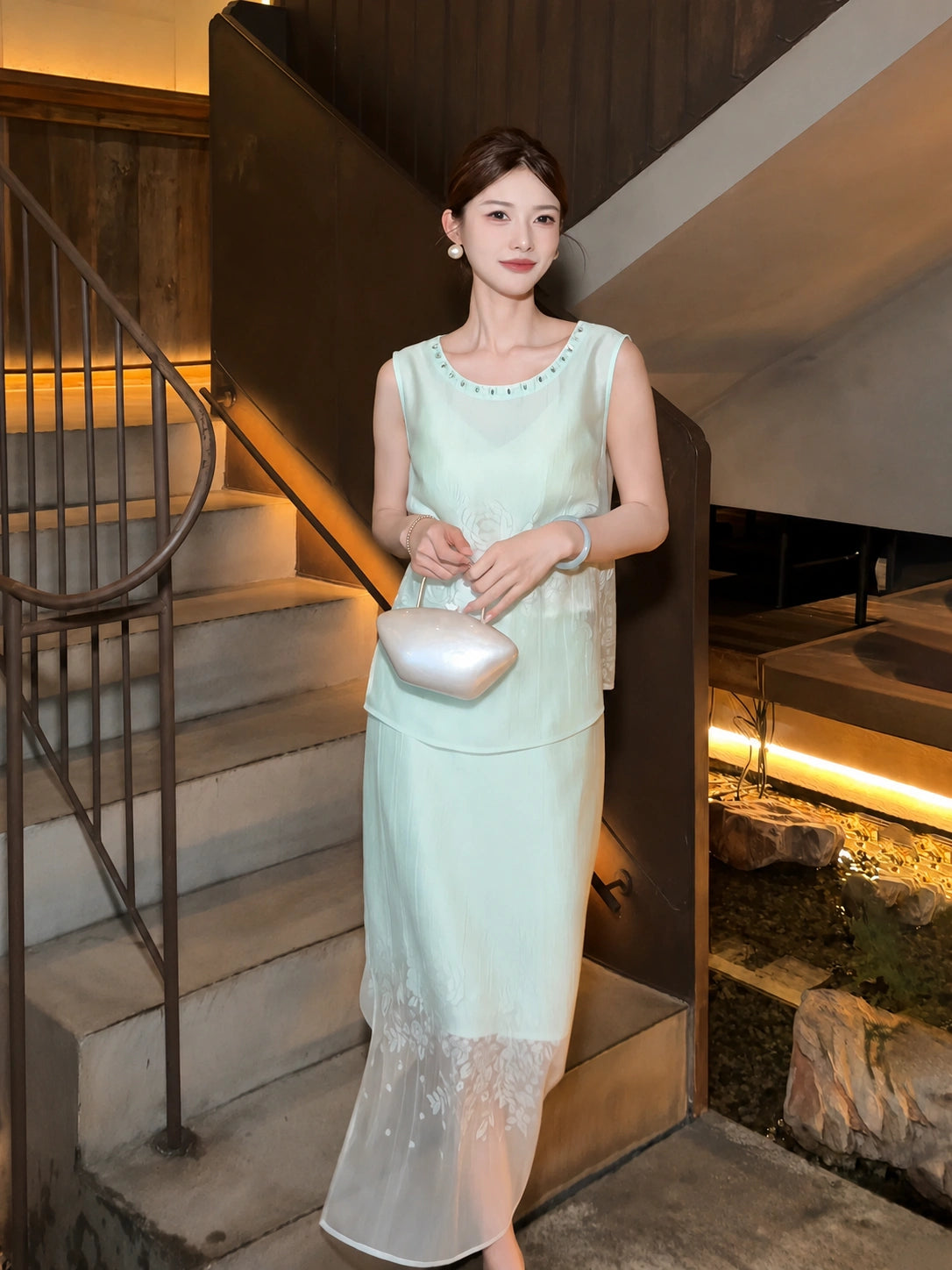 Model wearing a mint green sleeveless top and matching skirt with beaded neckline, standing on stairs, holding a white handbag.