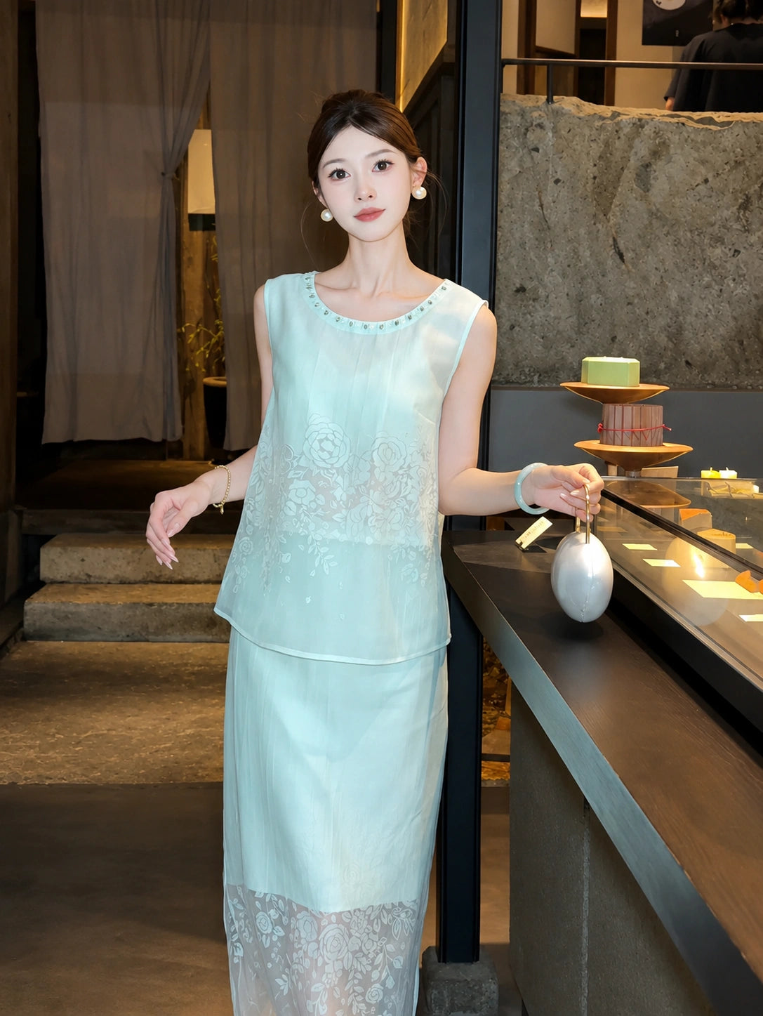 Model standing by a display case wearing a mint green embroidered set with a beaded round neckline, paired with a white handbag.