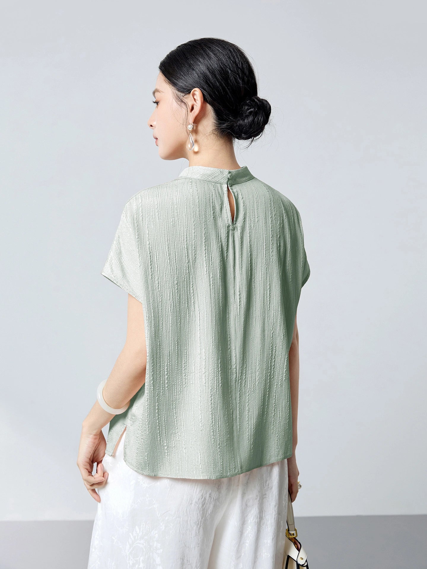 Back view of a mint green silk blouse featuring a stand collar, a back keyhole button closure, and a relaxed silhouette, paired with white pants.
