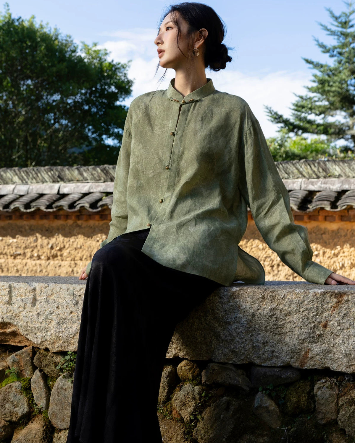 Model seated on a stone wall wearing a muted green stand-collar shirt with small front buttons and relaxed long sleeves.