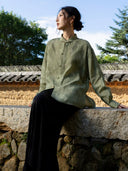 Model seated on a stone wall wearing a muted green stand-collar shirt with small front buttons and relaxed long sleeves.