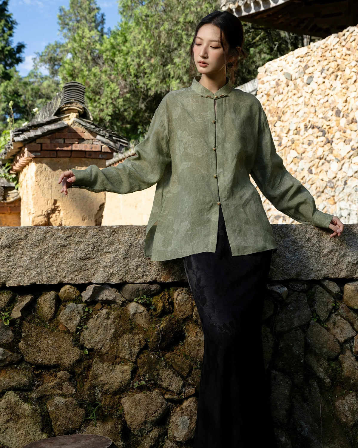 Model extending both arms to show the generous sleeves, relaxed shape and side slits of a muted green shirt.