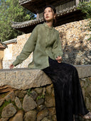 Model seated outdoors wearing a muted green shirt with a long black patterned skirt and metallic shoes.