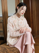 Seated model styling the embroidered porcelain white vest with a sheer blouse and pink skirt.