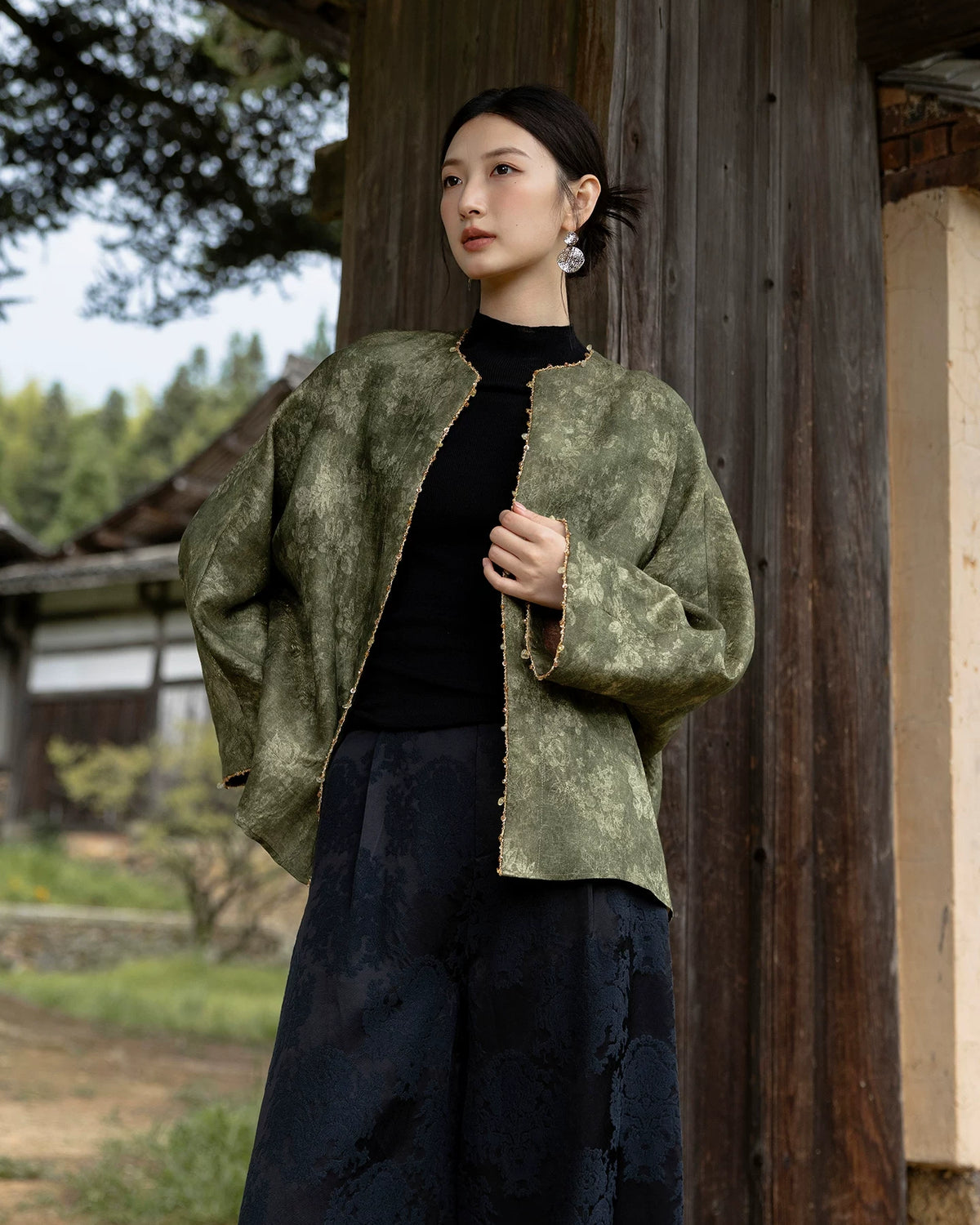 Model wearing an olive floral jacket with a round neckline and decorative gold-tone edging over a black top and dark trousers.