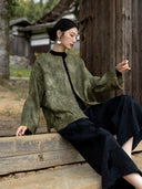 Model seated beside a wooden door, raising one arm to show the generous sleeve and trimmed cuff of an olive jacket.