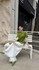 Model sitting on a white bench in an olive green short-sleeve Cheongsam top, crossing her legs, wearing white wide-leg pants and green shoes.