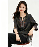 Sitting model wearing a dark green V-neck top with intricate red, blue, and white embroidery on the chest and sleeves, featuring a relaxed fit and 100% silk fabric.