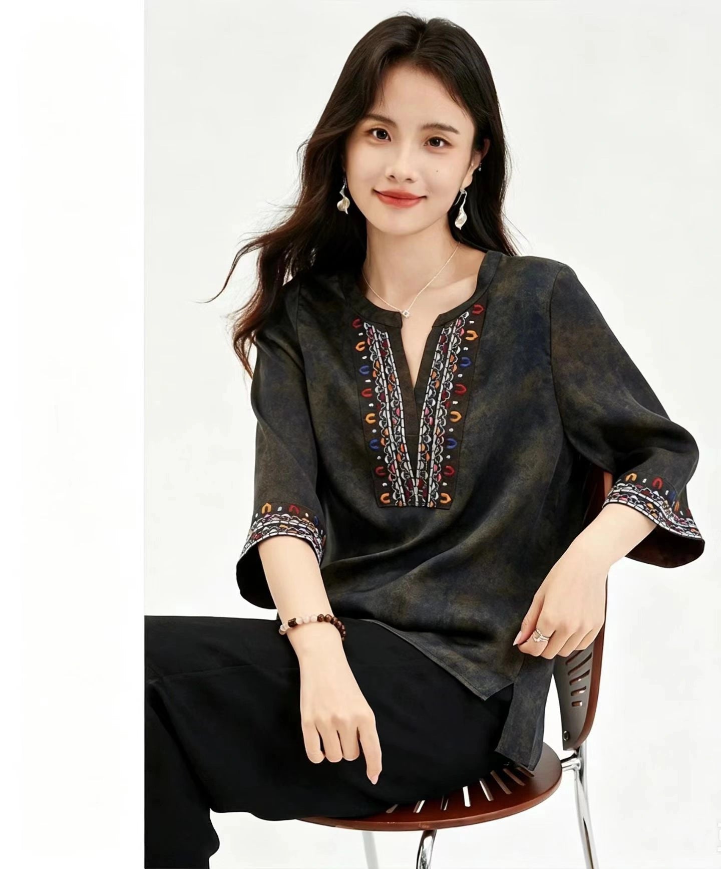 Sitting model wearing a dark green V-neck top with intricate red, blue, and white embroidery on the chest and sleeves, featuring a relaxed fit and 100% silk fabric.