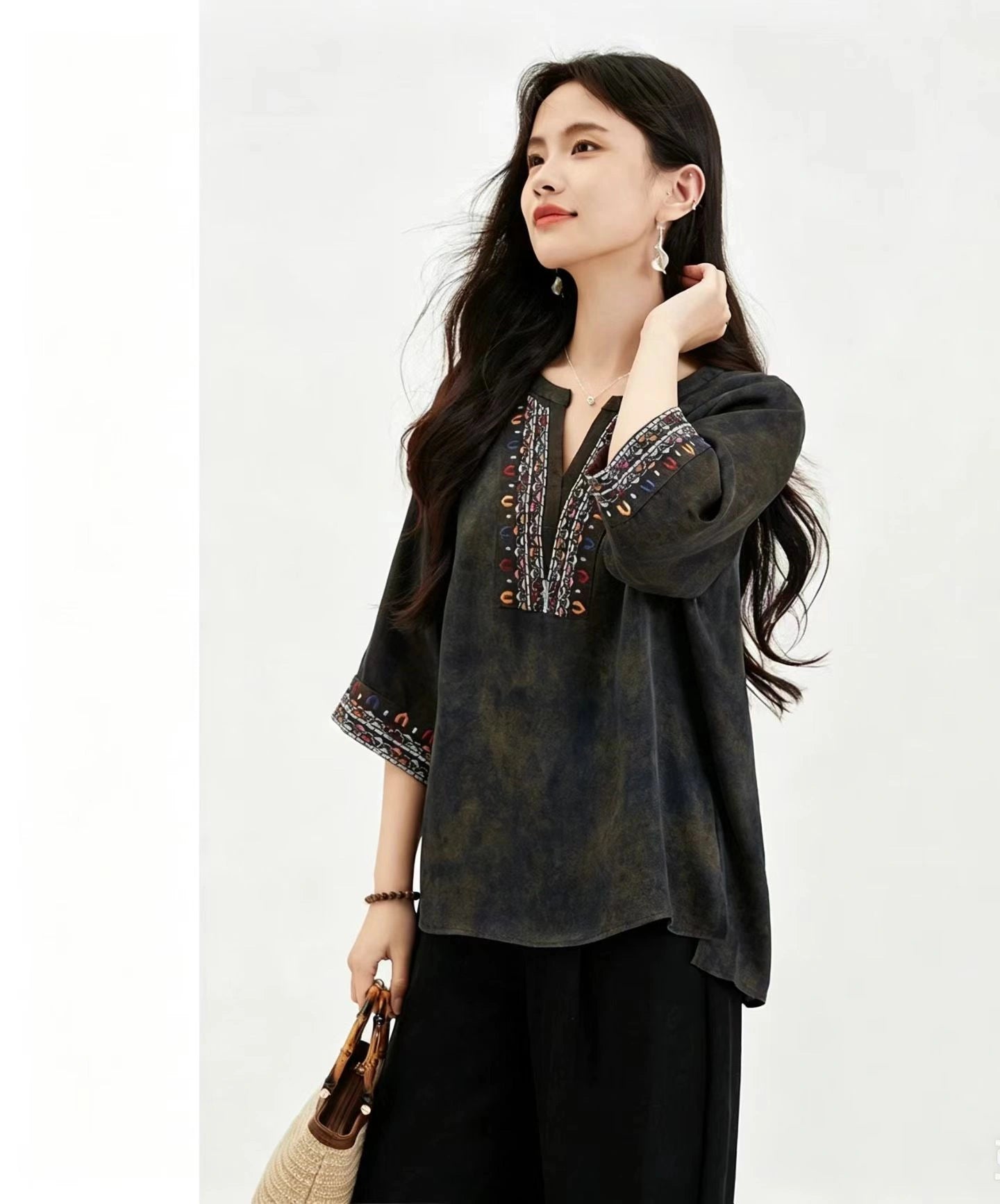 Full body shot of a women's olive green short sleeve silk top with embroidered details, styled with black trousers and a woven bag, highlighting the comfortable and elegant silhouette.