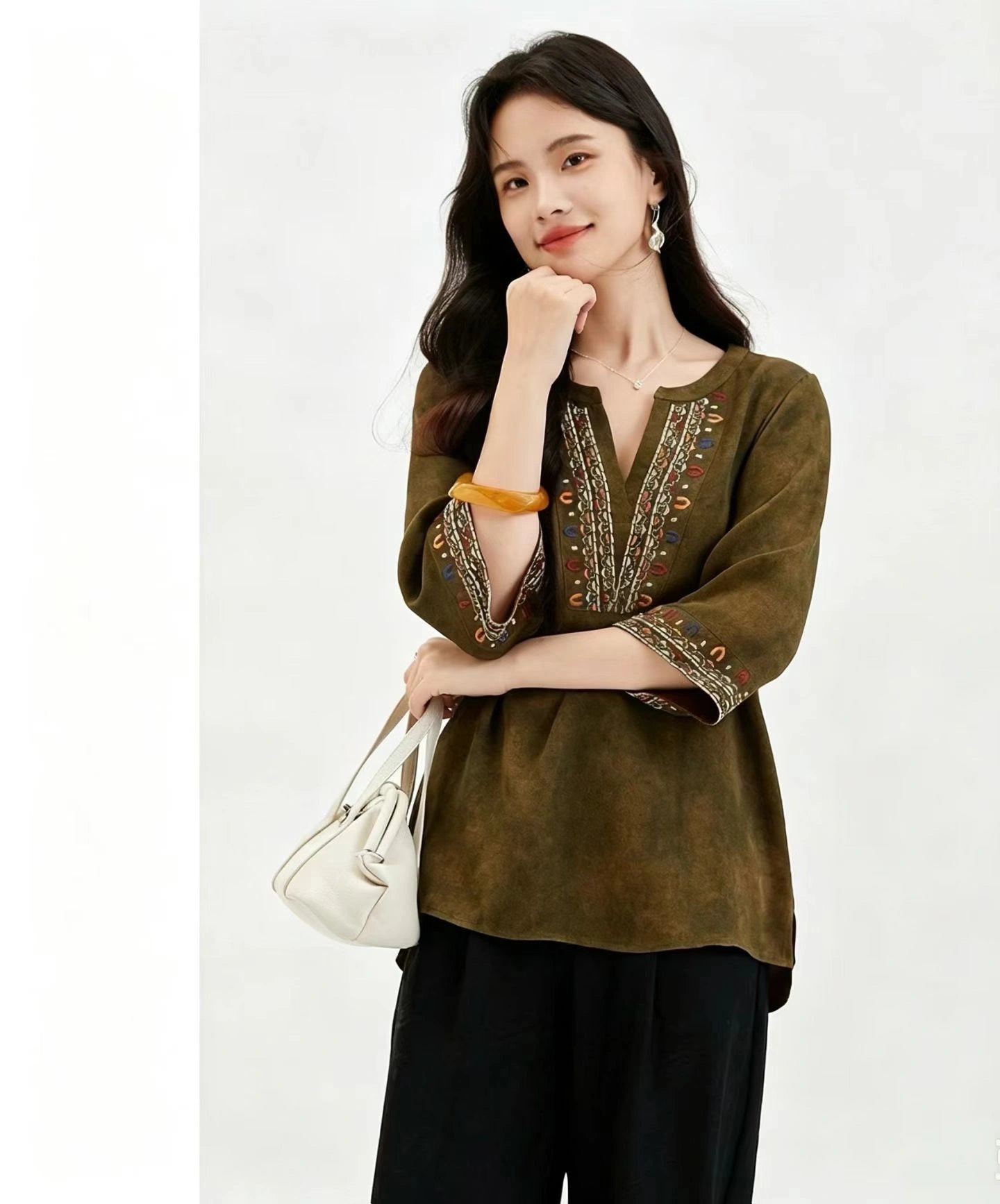 Front view of a deep olive green silk blouse with a V-neckline and colorful embroidery along the neck and cuffs, crafted from 100% mulberry silk, paired with black pants and a white handbag.
