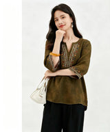Front view of a deep olive green silk blouse with a V-neckline and colorful embroidery along the neck and cuffs, crafted from 100% mulberry silk, paired with black pants and a white handbag.