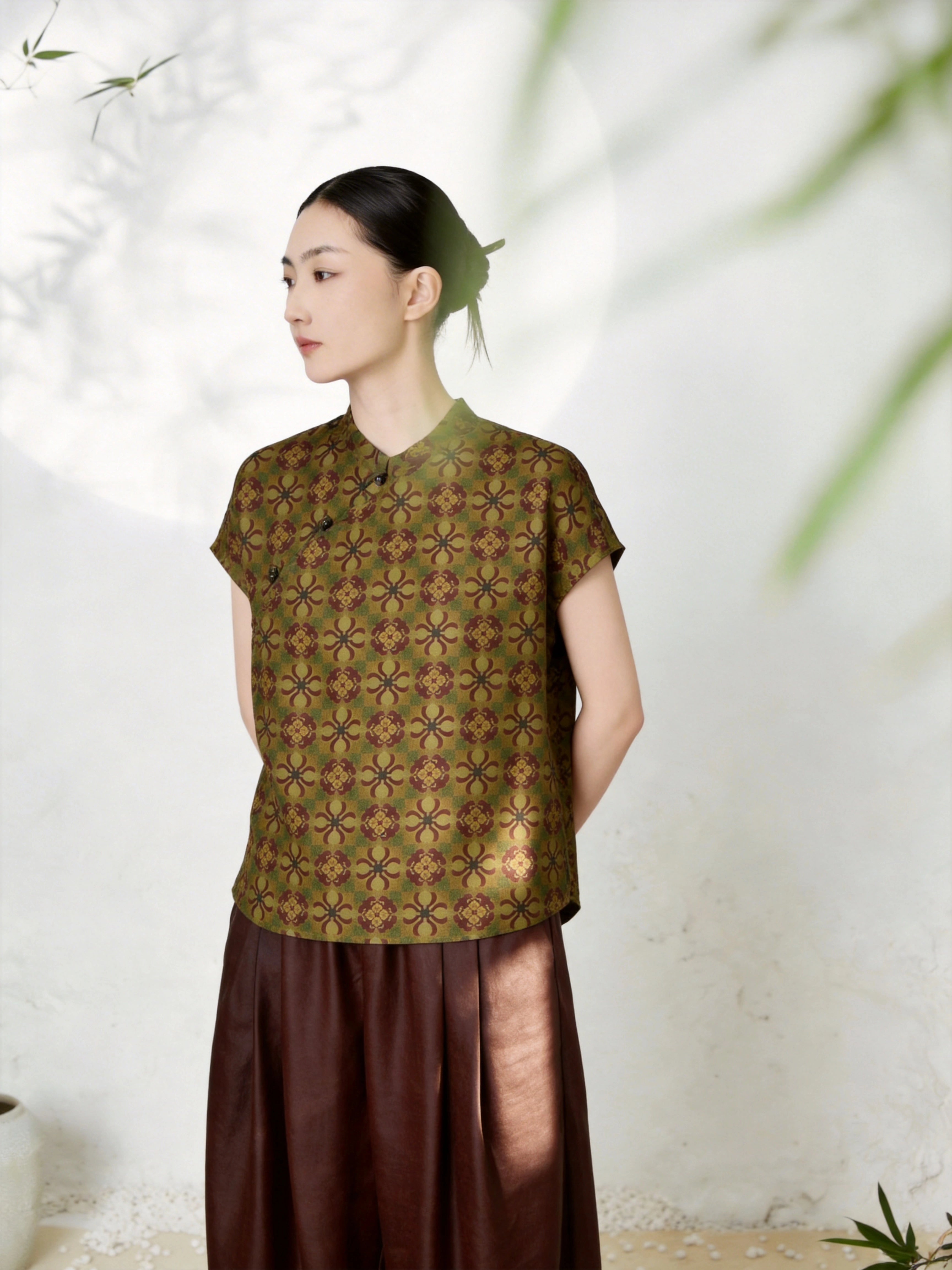 Side view of an olive green silk Cheongsam top with a mandarin collar, short sleeves, and all-over geometric jacquard pattern, featuring traditional side button details, crafted from 100% mulberry silk, paired with a brown pleated skirt.