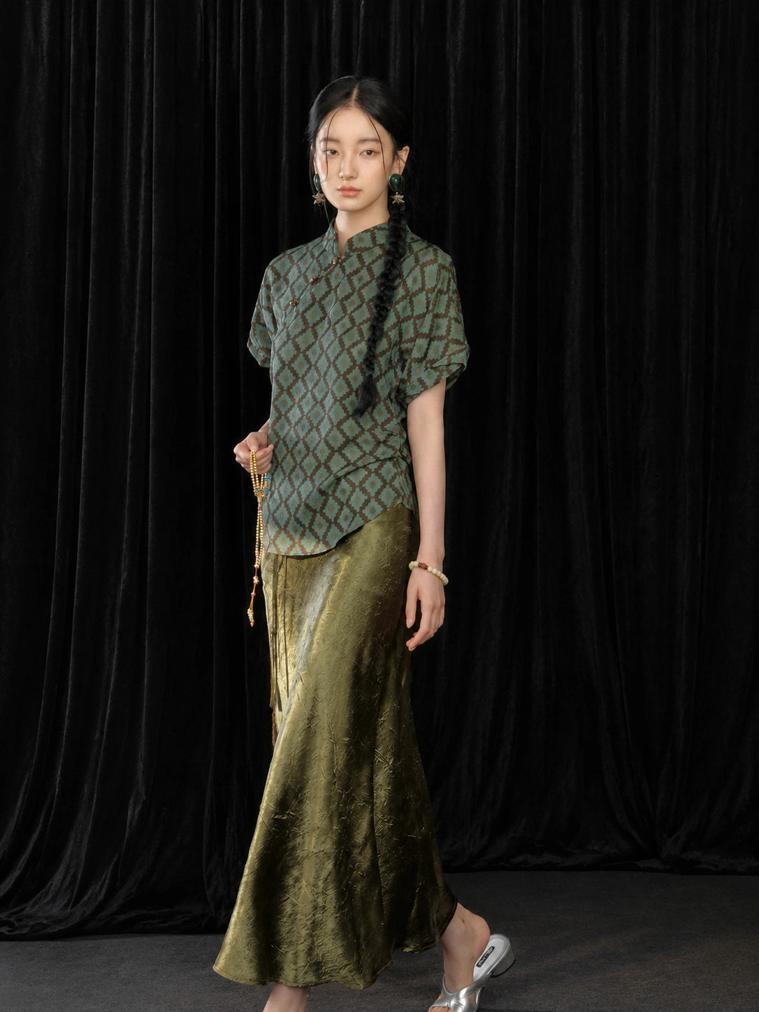 Model wearing an olive green high-sheen pleated fishtail skirt with a Chinese-style patterned top