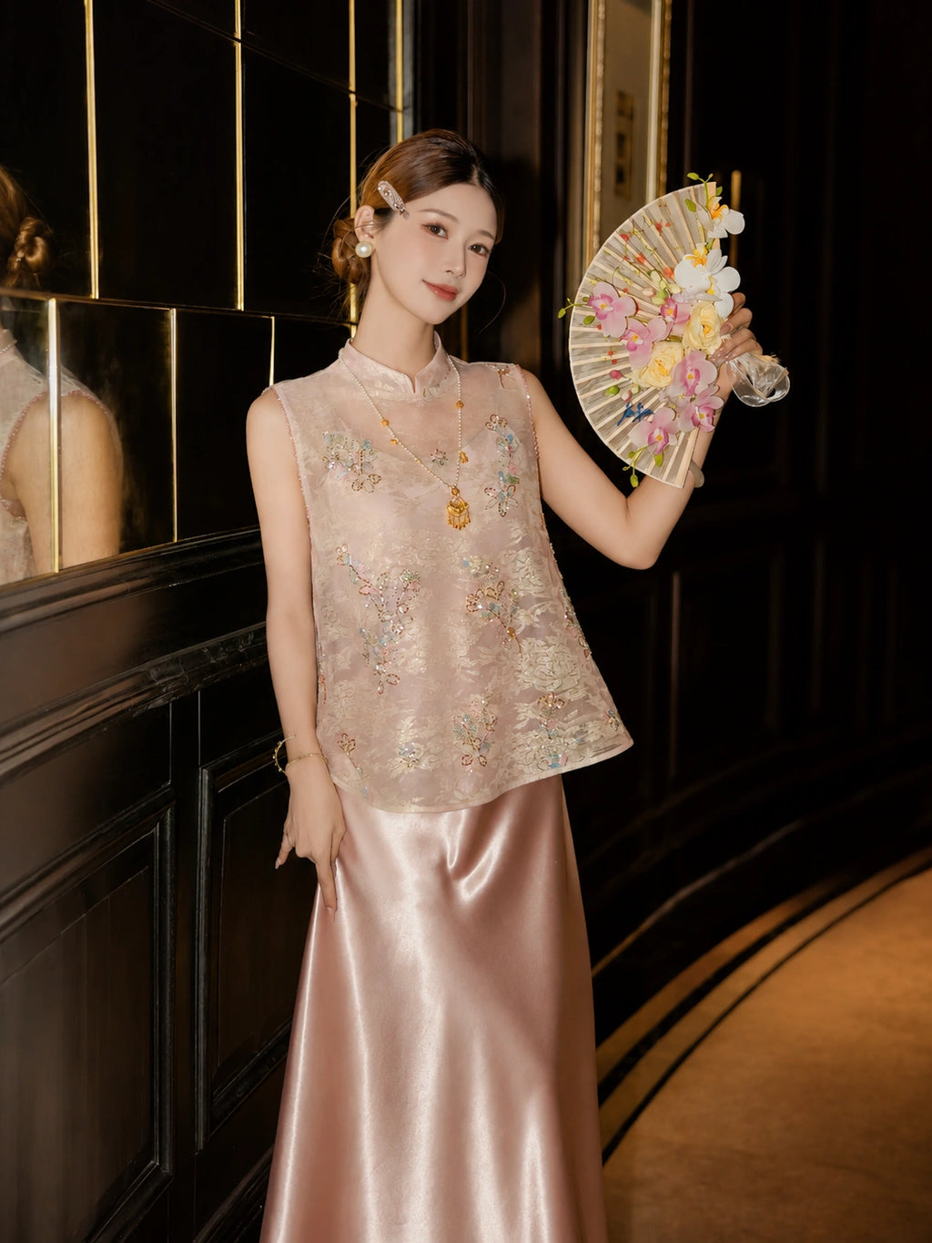 Model wearing a pink beaded Cheongsam vest with a mandarin collar and floral sequin details, holding a decorative fan, styled with a pink satin skirt.