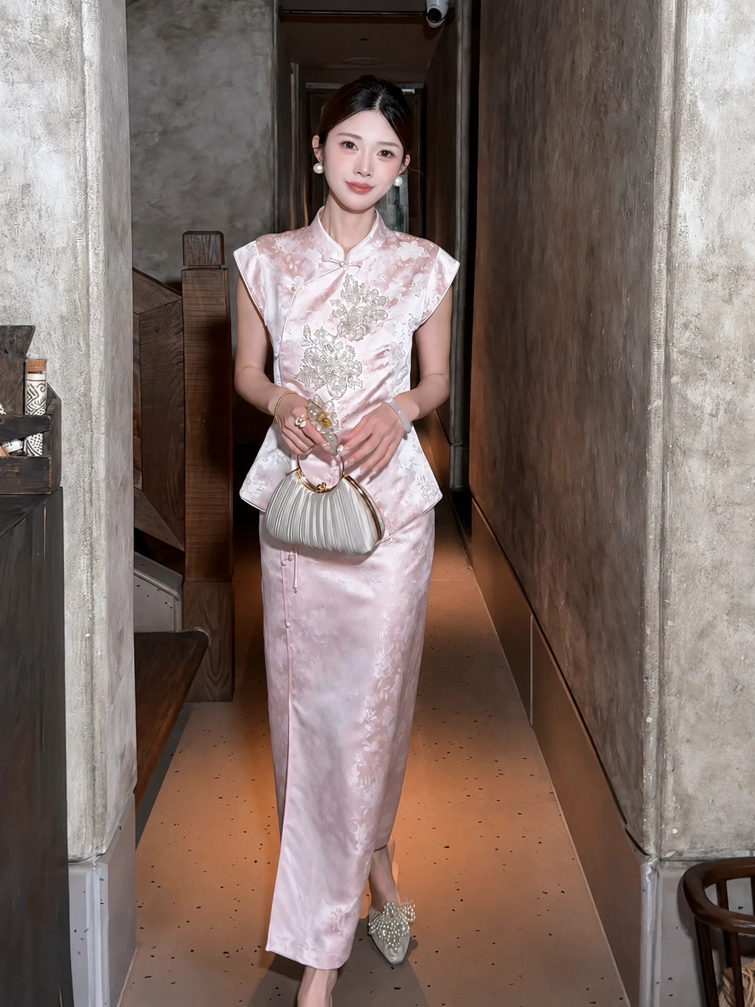 Full body shot of a pink Cheongsam set with cap sleeves and floral embroidery, styled with pearl shoes and a gold handbag, walking down a hallway.