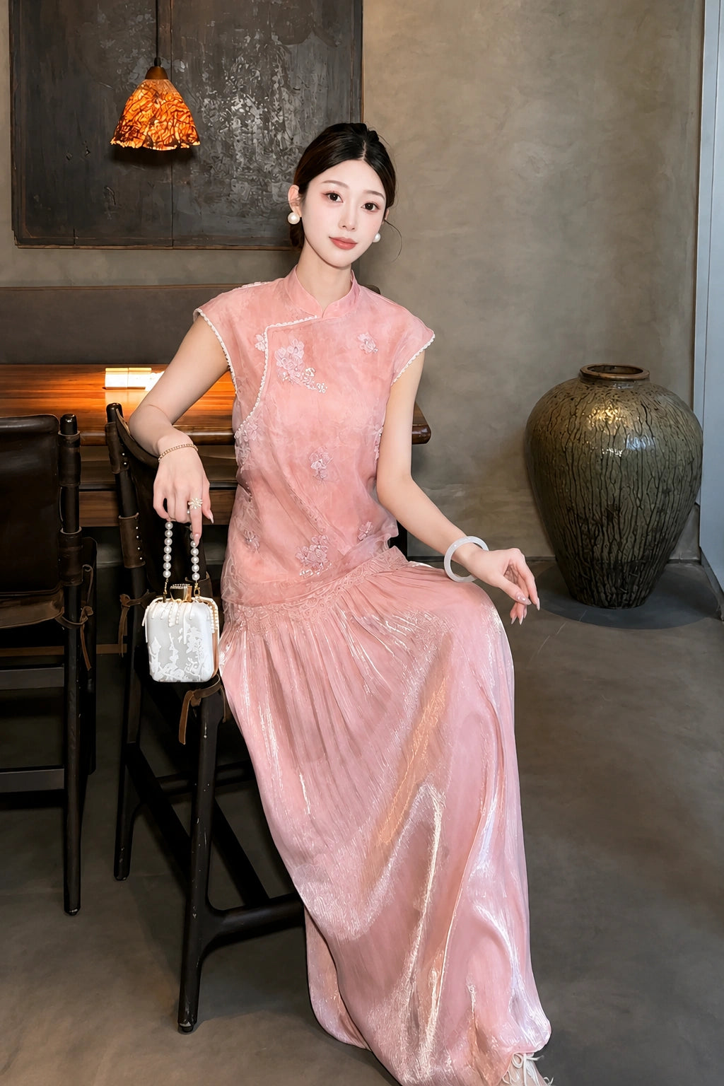 Sitting model in a pink Cheongsam dress with a mandarin collar, delicate embroidery, and a pleated skirt, holding a small bag, showcasing the relaxed fit.