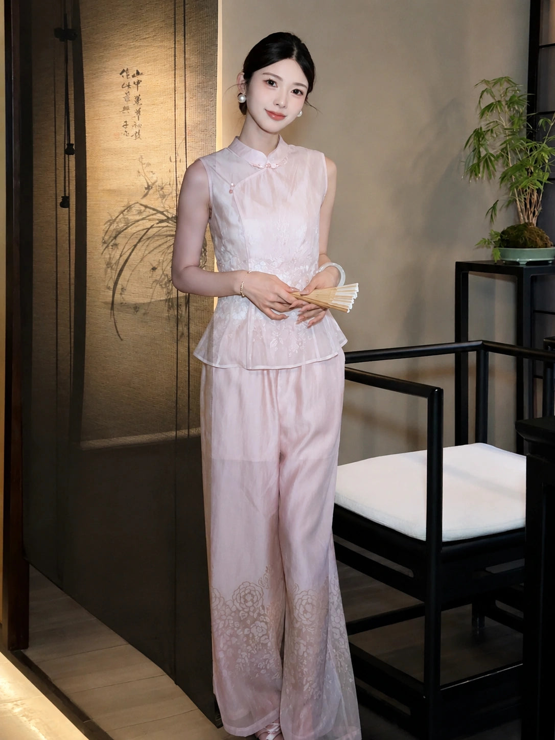 Women's pink sleeveless Qipao top and wide-leg pants set with textured fabric, styled with a paper fan in a traditional setting.