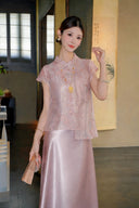 Model wearing a pink short-sleeve embroidered top with a mandarin collar and side ties, paired with a matching satin skirt, holding a fan.