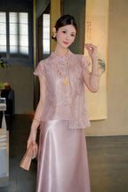 Model wearing a pink short-sleeve embroidered top with a mandarin collar and side ties, paired with a matching satin skirt, holding a fan.