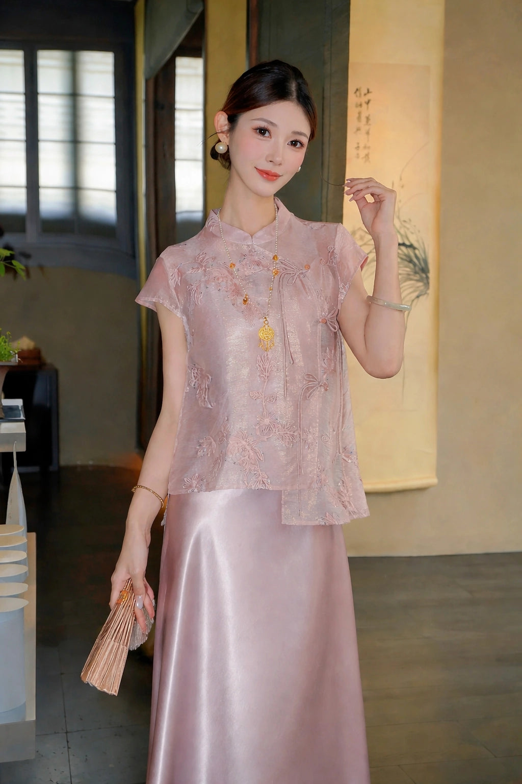 Model wearing a pink short-sleeve embroidered top with a mandarin collar and side ties, paired with a matching satin skirt, holding a fan.