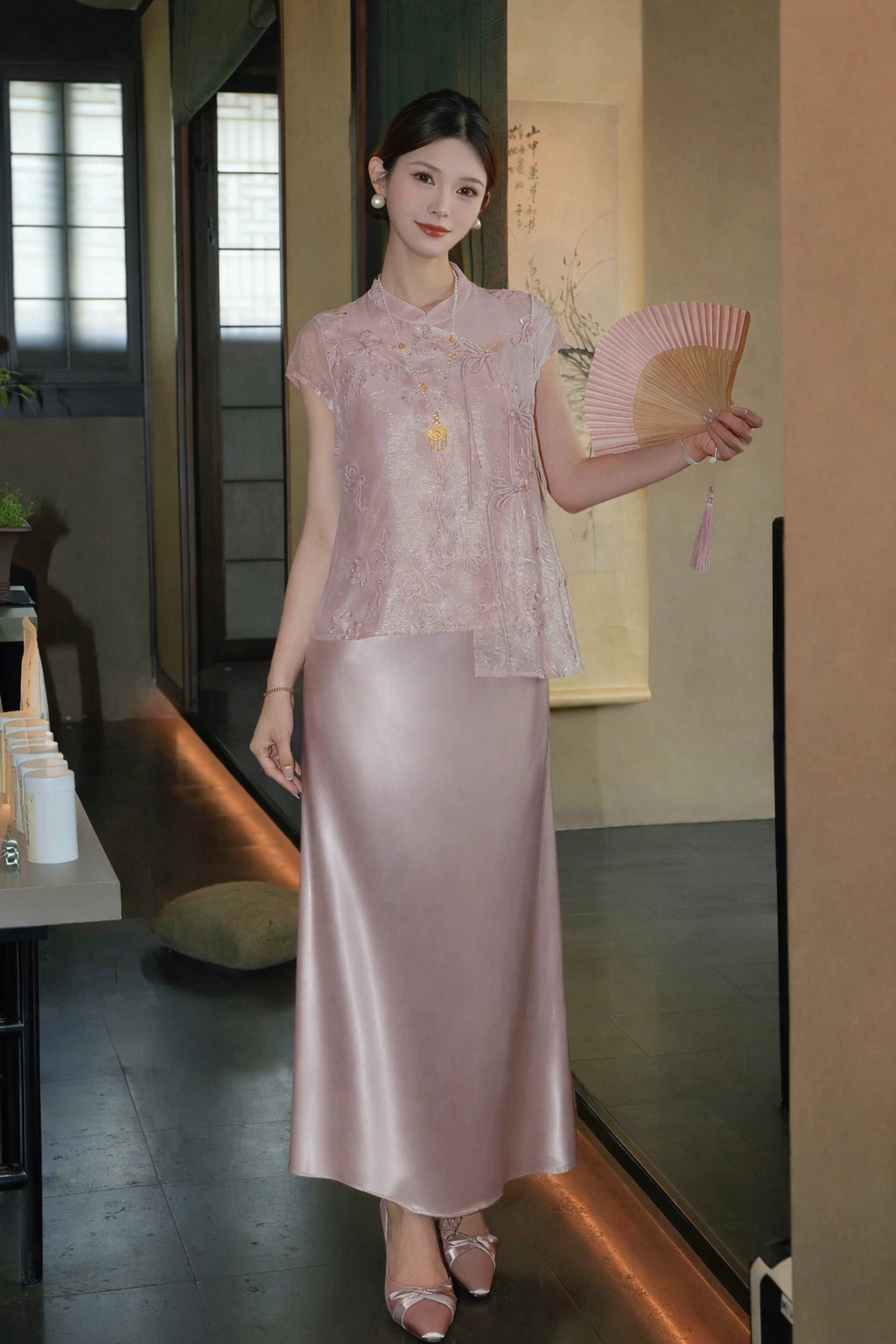 Full body shot of a pink short-sleeve Cheongsam top with floral embroidery and side ties, styled with a matching satin skirt and a gold necklace.