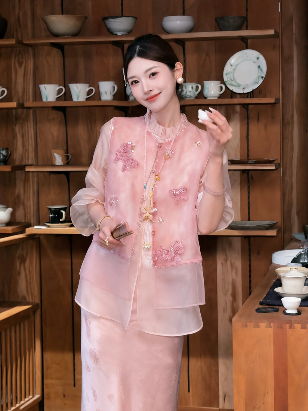 Model wearing a pink sleeveless embroidered vest with beadwork, styled over a sheer blouse and pink skirt, holding a small object in a tea room setting.