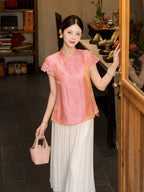Full body shot of a pink jacquard blouse with lace cap sleeves and a mandarin collar, styled with white pleated pants and a pink handbag.