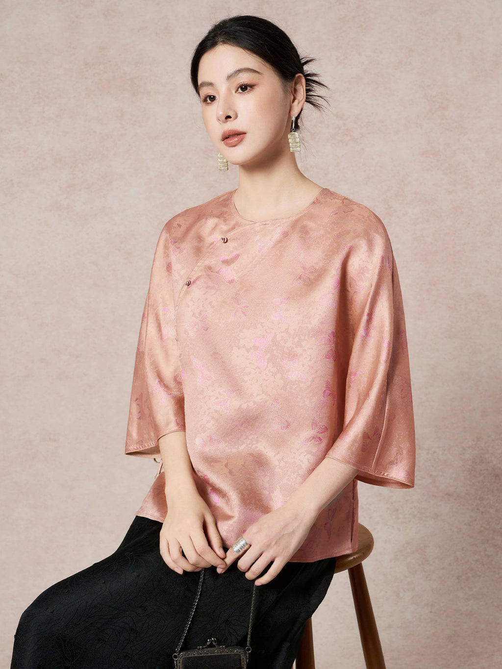 Model wearing a pink-gold silk shirt with a butterfly jacquard pattern, sitting and holding a bag, highlighting the elegant drape and comfortable fit.