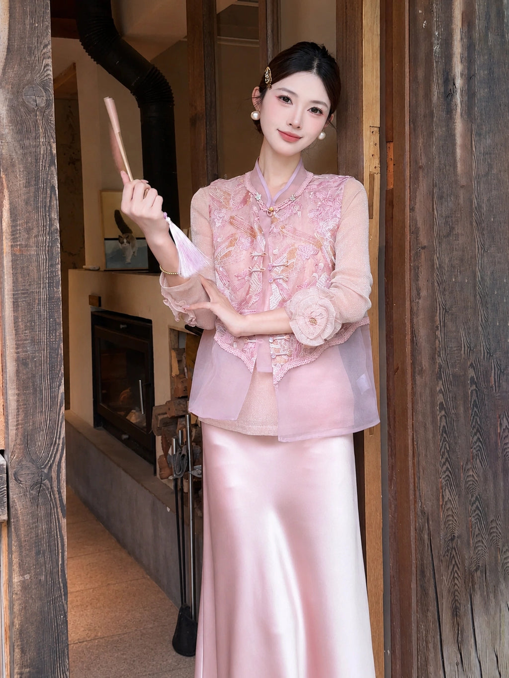 Model wearing a pink sleeveless embroidered vest with a mandarin collar and gold buttons, paired with a pink satin skirt, holding a fan.