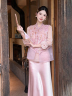 Model wearing a pink sleeveless embroidered vest with a mandarin collar and gold buttons, paired with a pink satin skirt, holding a fan.