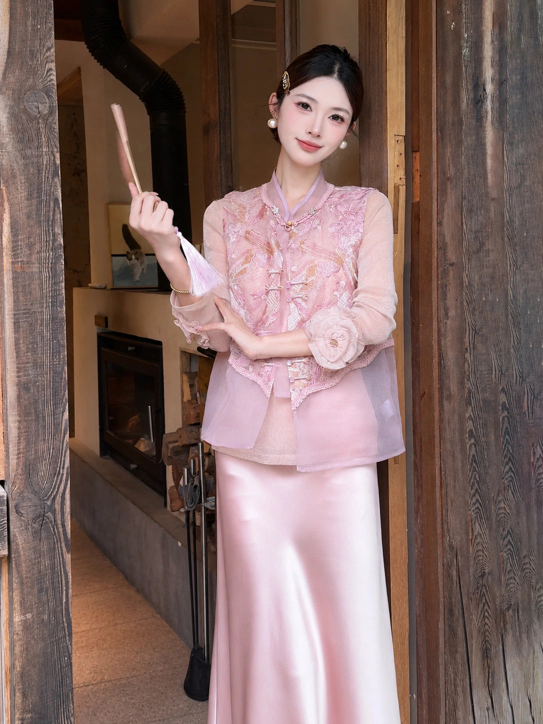 Model wearing a pink sleeveless embroidered vest with a mandarin collar and gold buttons, paired with a pink satin skirt, holding a fan.