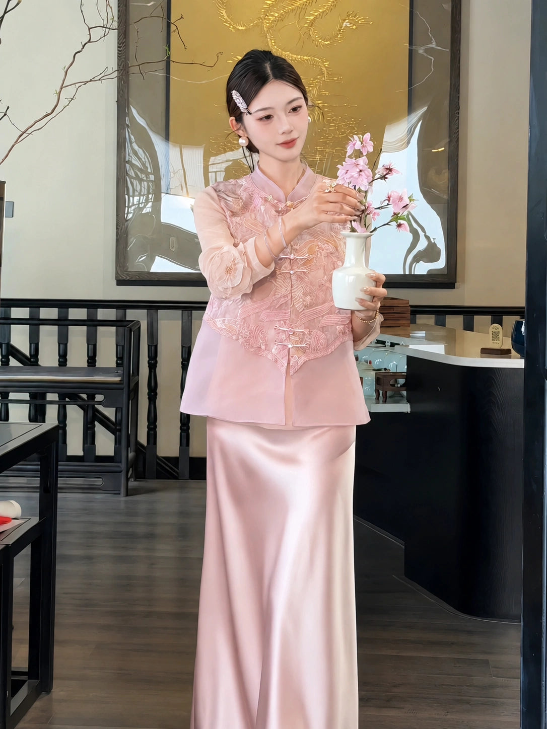 Front view of a pink sleeveless vest with metallic embroidery and sheer sleeves, styled with a pink satin skirt, holding a white vase with flowers.