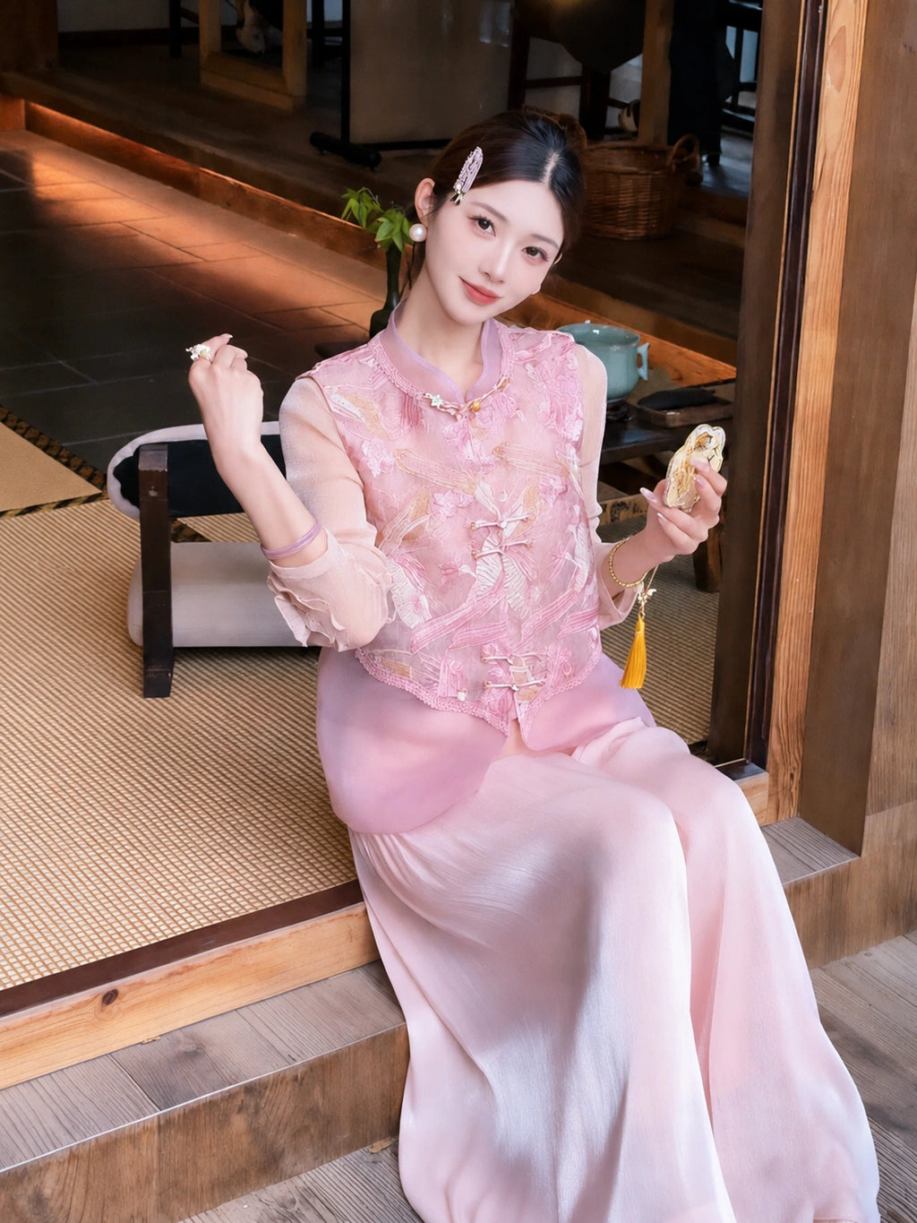 Sitting model in a pink embroidered vest with gold buttons and sheer layered hem, holding a fan, showcasing the relaxed fit and elegant drape.