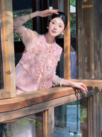 Model leaning on a wooden window frame wearing a pink sleeveless vest with metallic embroidery and sheer sleeves, styled with a pink skirt.