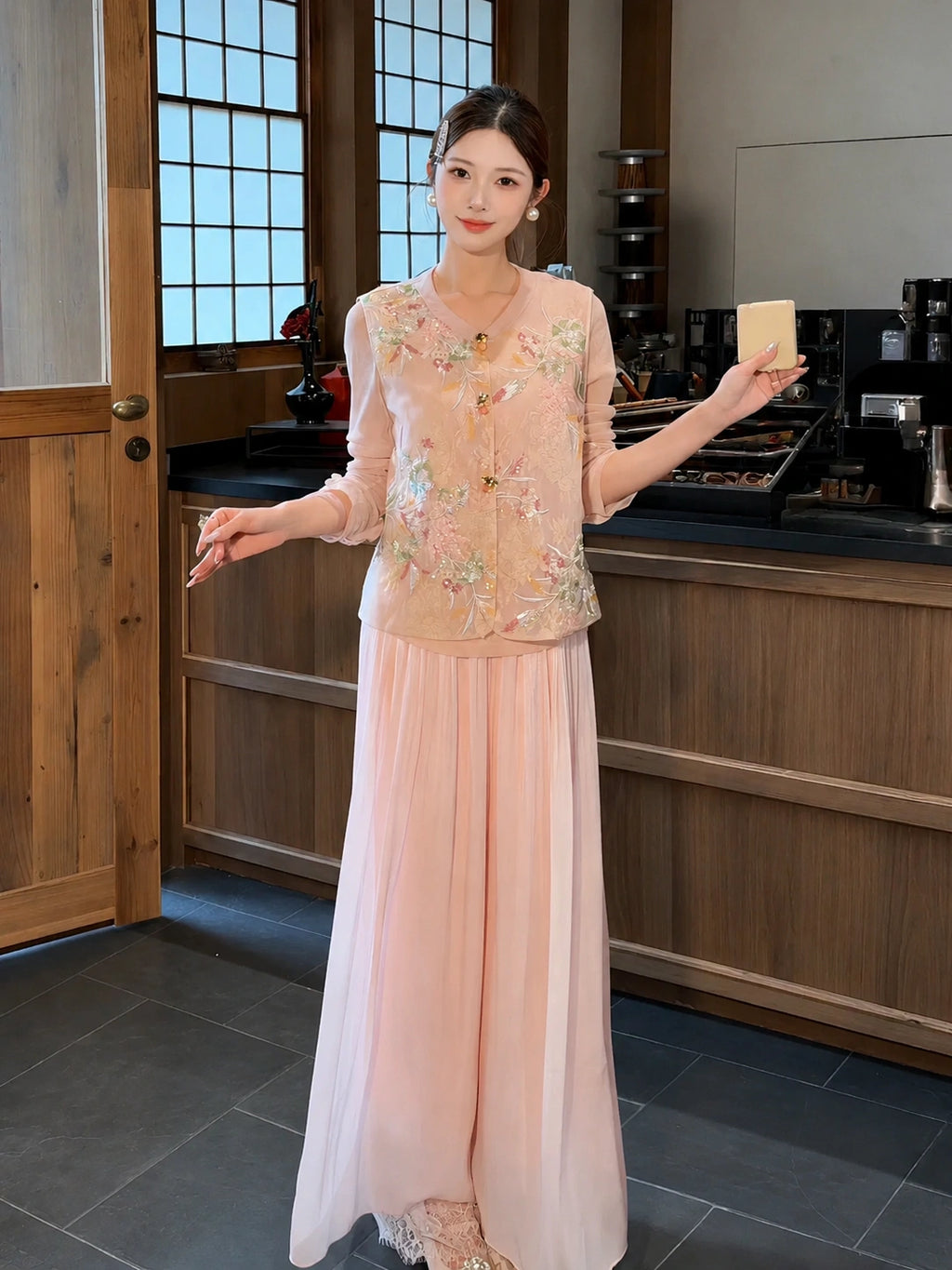 Model wearing pink pleated wide-leg pants with a layered lace hem, paired with an embroidered vest, holding a small object, showcasing the flowy silhouette.