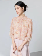 Model seated wearing a pink floral 100% mulberry silk blouse with a ruffled collar, front pleats, and puff sleeves, styled with a matching skirt.
