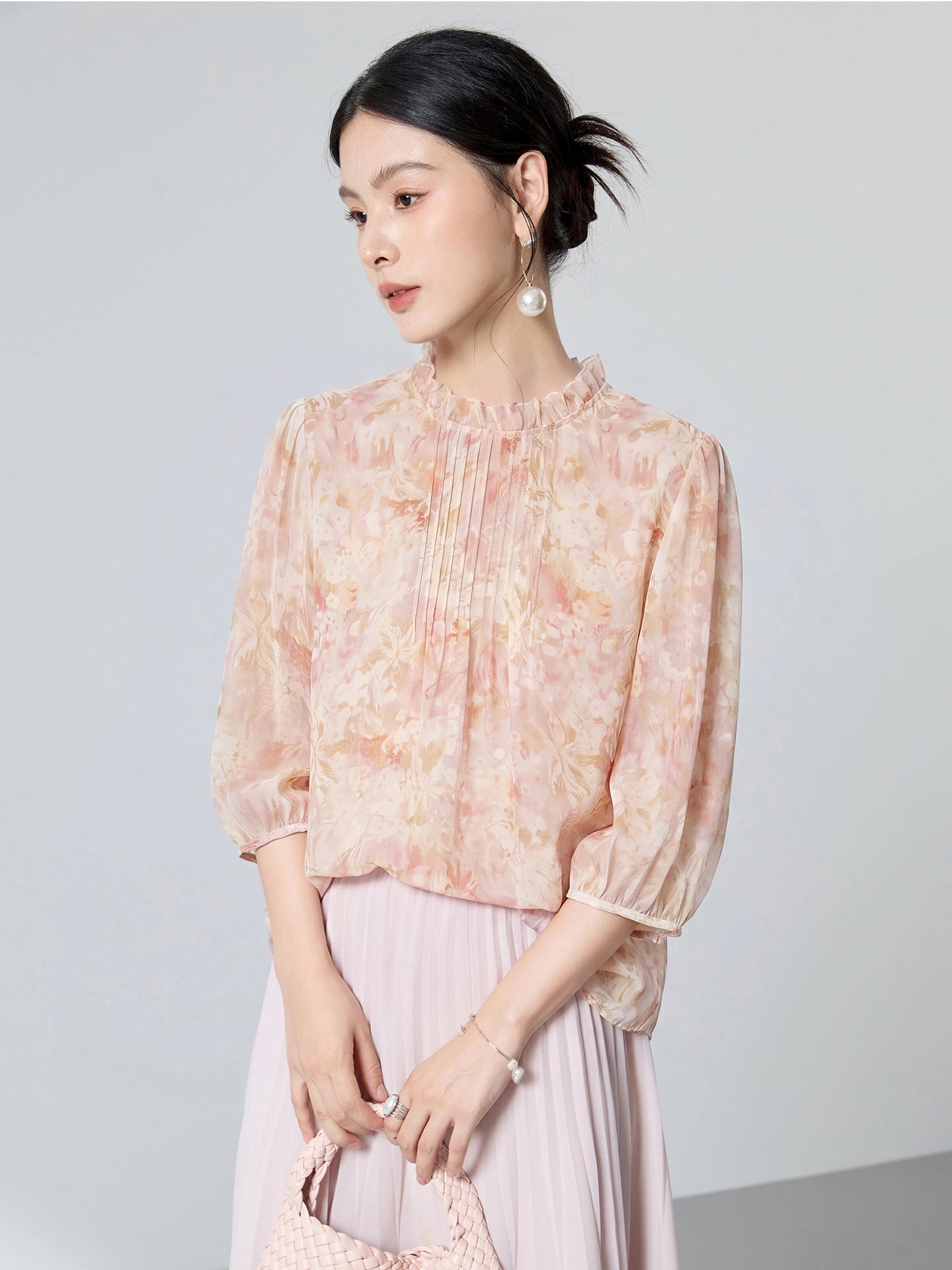 Model seated wearing a pink floral 100% mulberry silk blouse with a ruffled collar, front pleats, and puff sleeves, styled with a matching skirt.