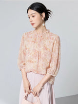 Model seated wearing a pink floral 100% mulberry silk blouse with a ruffled collar, front pleats, and puff sleeves, styled with a matching skirt.