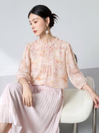 Model seated wearing a pink floral 100% mulberry silk blouse with a ruffled collar, front pleats, and puff sleeves, styled with a matching skirt.